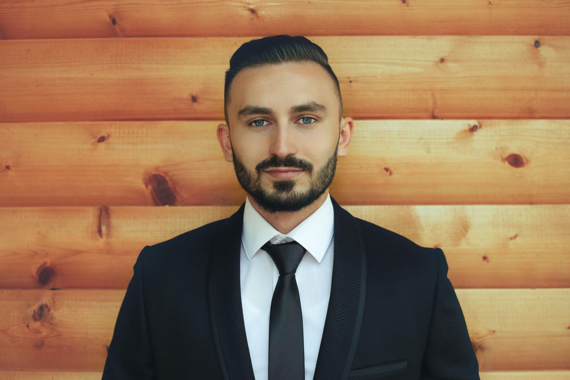 Man with a beard in a suit and tie standing against a wooden plank wall.